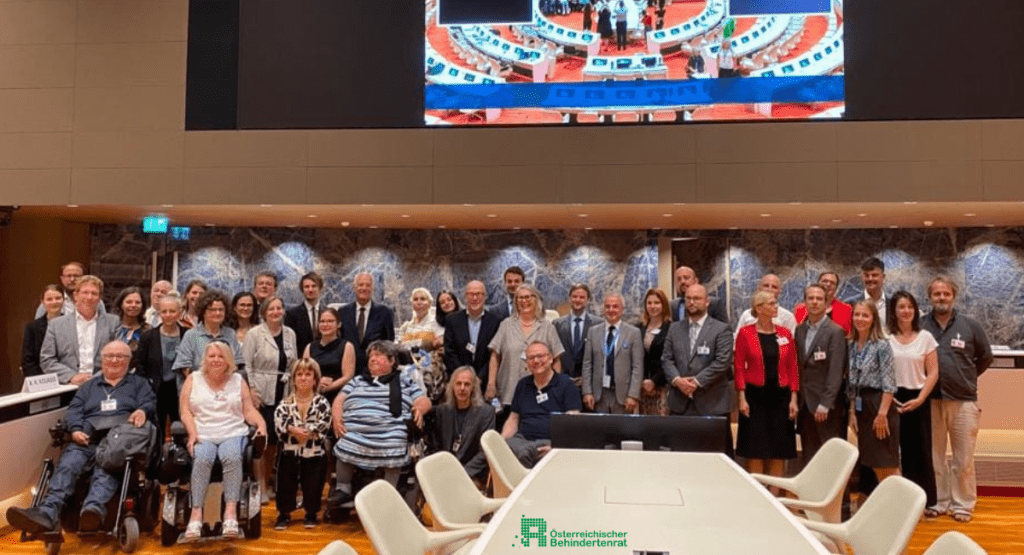 Bernadette Feuerstein mit einer Delegation in einem großen Konferenzsaal. Die Gruppe posiert für ein gemeinsames Foto, einige Personen sitzen im Rollstuhl.