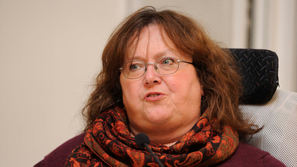 Bernadette Feuerstein spricht vor einem Mikrofon. Sie trägt eine Brille und einen rot gemusterten Schal.