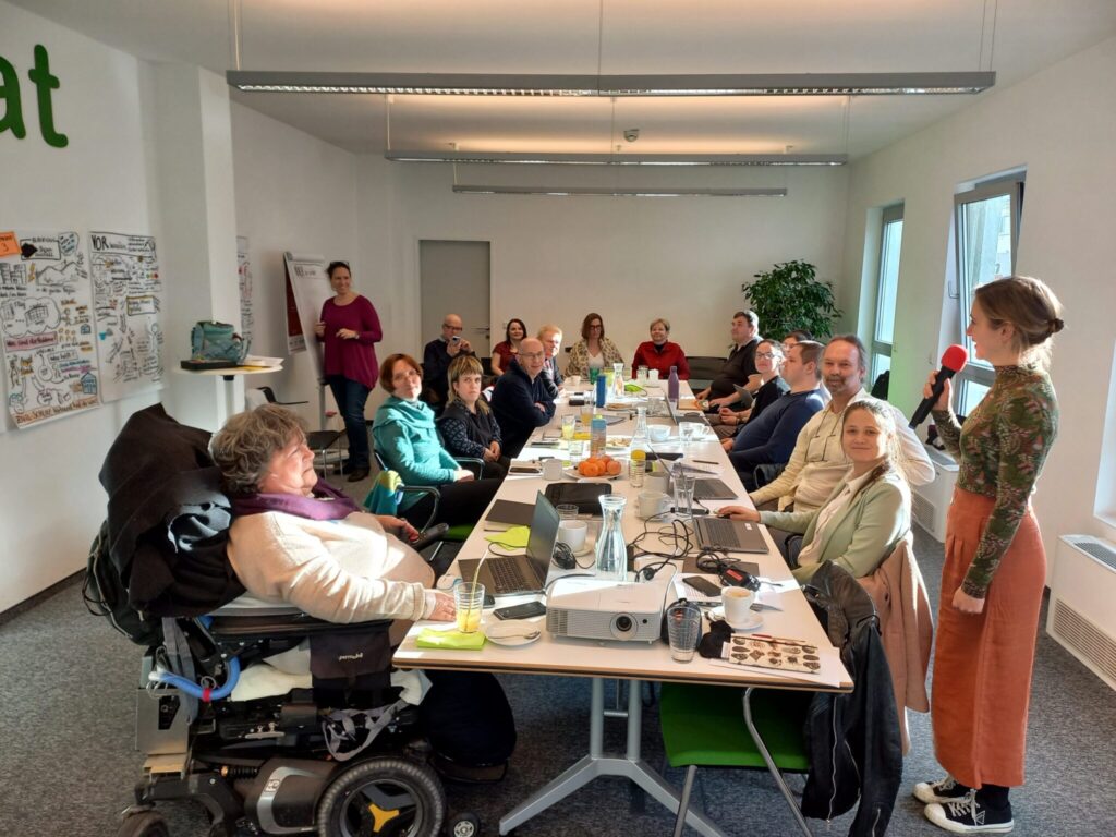 Bernadette Feuerstein sitzt im Elektrorollstuhl an einem Konferenztisch. Mehrere Personen nehmen an einem Workshop teil, während eine Frau mit Mikrofon spricht.