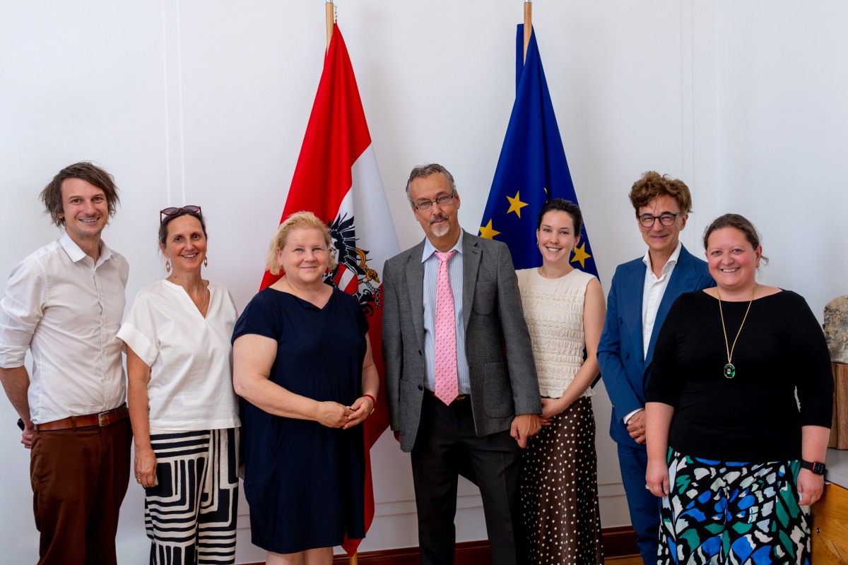 Gruppenfoto eines Treffens zwischen Vertreterinnen des Sozialministeriums und des Österreichischen Behindertenrats. Sieben Personen stehen nebeneinander vor einer weißen Wand, zwischen einer österreichischen Staatsflagge und einer EU-Flagge. In der Mitte stehen Sozialministerin Korinna Schumann und Behindertenrats-Präsident Klaus Widl. Die übrigen Teilnehmerinnen sind links und rechts davon angeordnet. Alle sind in Business- bzw. Business-Casual-Kleidung gekleidet und lächeln in die Kamera.