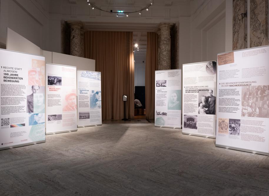 Ausstellung mit mehreren aufgestellten Informationstafeln in einem großen Raum mit hohen Säulen und Vorhang im Hintergrund