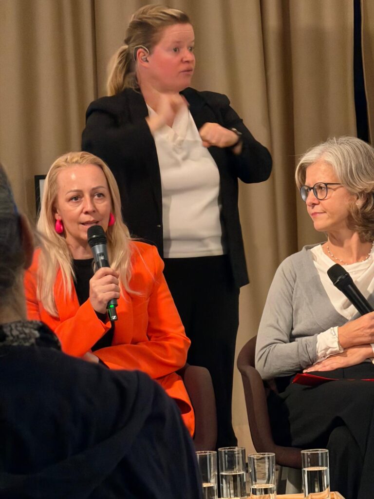 Christine Steger spricht bei einer Podiumsdiskussion in ein Mikrofon. Neben ihr sitzt Angela Wegscheider, im Hintergrund übersetzt eine Gebärdensprachdolmetscherin.
