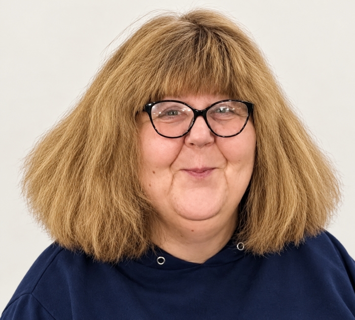 Porträt einer lächelnden Frau mit schulterlangem, blondem Haar und schwarzer Brille vor hellem Hintergrund.