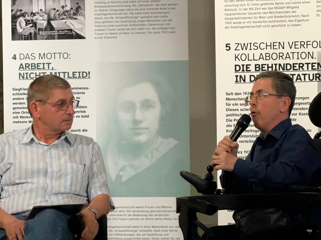 Kurt G. Beck (links) und Martin Ladstätter im Gespräch vor Ausstellungstafeln. Ladstätter sitzt im Rollstuhl und spricht in ein Mikrofon.