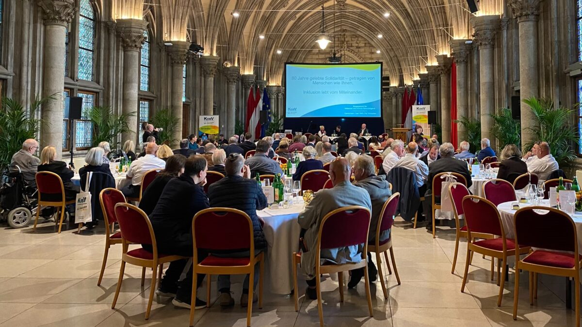 Zahlreiche Gäste sitzen an festlich gedeckten runden Tischen in der Volkshalle des Wiener Rathauses anlässlich des 80-jährigen Jubiläums des KOBV. Der historische Saal ist von hohen Säulen, einem Kreuzrippengewölbe und großen bunten Glasfenstern geprägt.