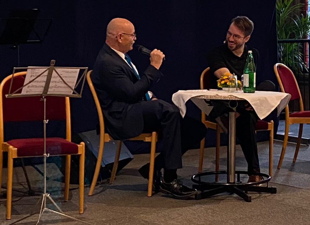 KOBV-Präsident Franz Groschan (links) spricht mit Moderator Andreas Onea (rechts) bei der Feier zum 80-jährigen Bestehen des KOBV im Wiener Rathaus. Beide sitzen auf der Bühne an einem kleinen Tisch und sehen einander lächelnd an. Groschan hält ein Mikrofon in der Hand.