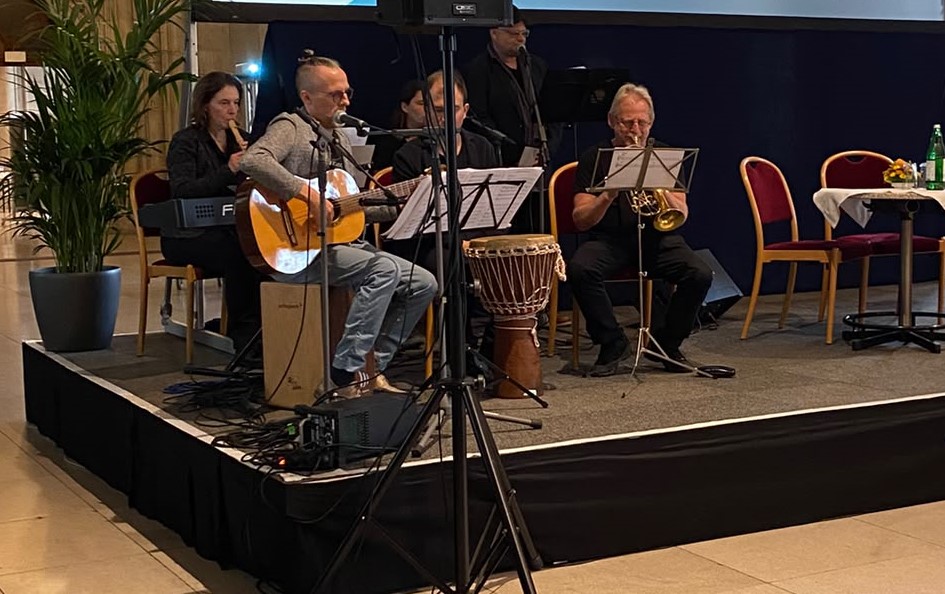 Das inklusive Ensemble Ohrenklang musiziert bei der 80-Jahr-Feier des KOBV auf einer Bühne. Ein Musiker spielt Gitarre und singt ins Mikrofon, weitere Ensemblemitglieder spielen unter anderem Trompete, Keyboard und Blasinstrumente. Vor ihnen stehen Notenständer und eine Trommel.