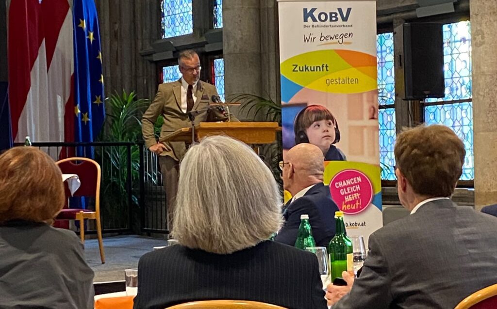 Behindertenrats-Präsident Klaus Widl hält beim Festakt zum 80-jährigen Jubiläum des KOBV im Wiener Rathaus eine Rede. Er steht im Anzug hinter einem Rednerpult, neben ihm ein KOBV-Roll-up. Im Vordergrund verfolgen mehrere Gäste seine Ansprache.