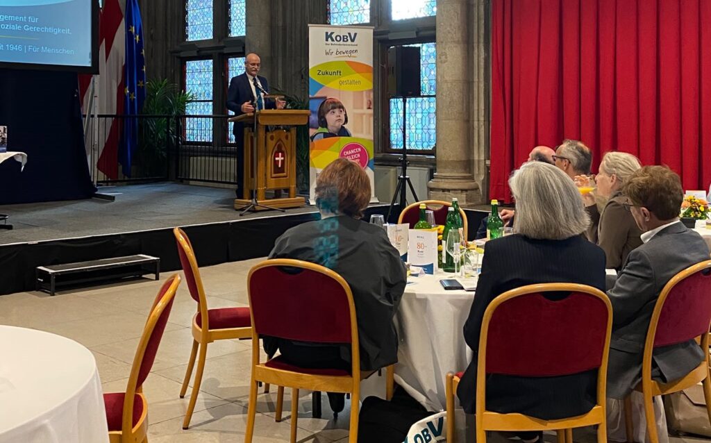 KOBV-Präsident Franz Groschan hält beim Festakt zum 80-jährigen Jubiläum des KOBV im Wiener Rathaus eine Rede. Er steht im Anzug hinter einem Rednerpult, neben ihm ein KOBV-Roll-up. Im Vordergrund verfolgen mehrere Gäste seine Ansprache.