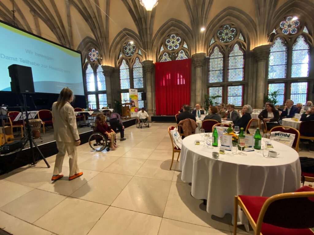 Mitglieder von DanceAbility Austria führen bei der 80-Jahr-Feier des KOBV in der Volkshalle des Wiener Rathauses eine inklusive Tanzperformance auf. Mehrere Tänzer:innen, darunter Personen im Rollstuhl, bewegen sich vor der Bühne. Die Gäste verfolgen die Aufführung von ihren Tischen aus. Im Hintergrund sind die hohen Glasfenster des historischen Saals zu sehen.