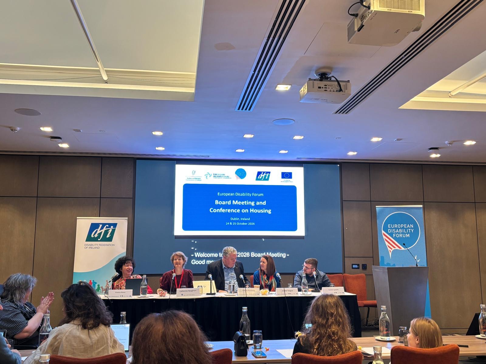 5 Personen sitzen an einem Podium und sprechen vor einem Publikum. Im Hintergrund der Titel: "Board Meeting and Conference on Housing".