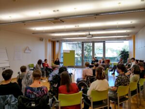 Viele Personen sitzen im Halbkreis, vorne sitzen 3 Frauen, die die Session leiten, eine davon ist im Rollstuhl.