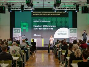 Eine Frau im Rollstuhl sitzt auf der Bühne. Hinter ihr steht "Herzlich Willkommen zur Fachkonferenz", rechts am Rand steht ein ÖGS Dolmetscher.