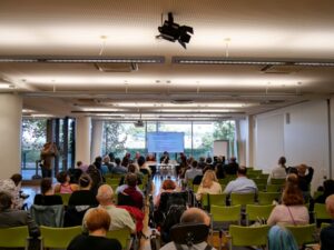 Viele Personen sitzen in einem Seminarraum, vorne sitzen 4 Personen die die Session leiten.