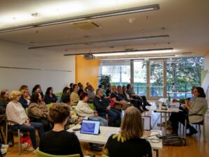 Viele Personen sitzen in einem Seminarraum, vor ihnen sitzen zwei Personen die die Session leiten. Am Rand sitzen zwei Schriftdolmetscherinnen.
