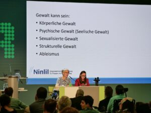Zwei Frauen sitzen auf der Bühne, eine spricht in ein Mikrofon. Hinter ihnen sind auf einer Powerpoint Folie verschiedene Gewaltformen erklärt.