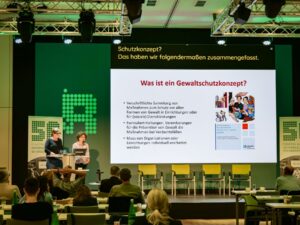 Zwei Frauen sprechen auf der Bühne, neben ihnen sieht man eine Powerpoint Präsentation, bei der Gewaltschutzkonzepte erklärt werden.