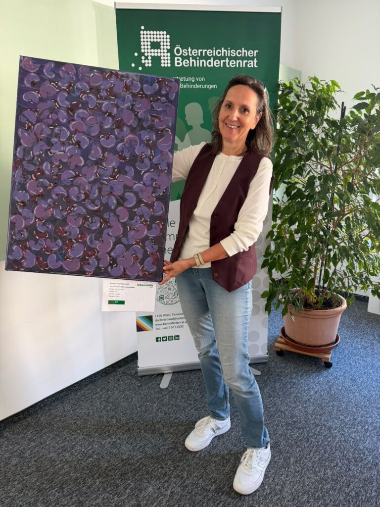 Barbara Haider-Novak steht neben einem Ficus und hält ein lila-bordeauxfarbenes Bild in Händen.