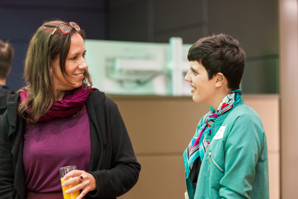 Petra Plicka und Nicole Osimk bei der Fachkonferenz des Österreichischen Behindertenrats 2026 unterhalten sich lächelnd. Plicka hält ein Glas Orangensaft in der Hand.