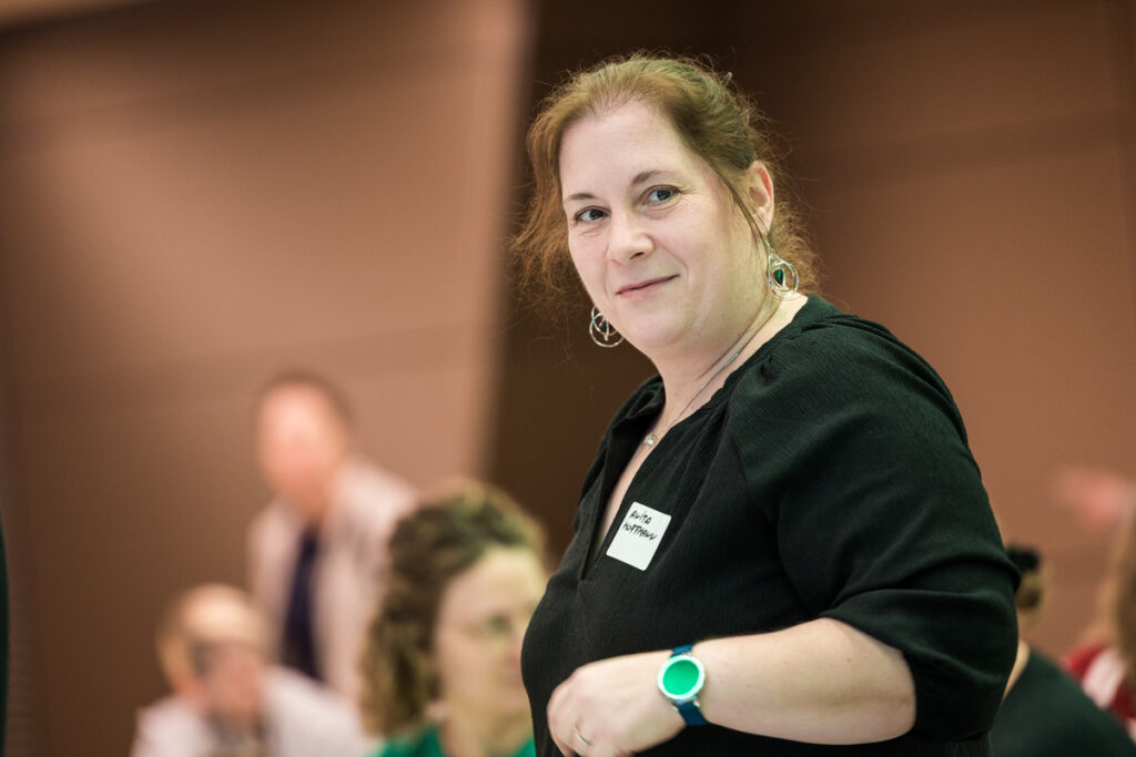 Anita Hoffmann, Teilnehmerin der Fachkonferenz des Österreichischen Behindertenrats 2026, blickt lächelnd in die Kamera. Sie trägt ein schwarzes Oberteil und eine grüne Armbanduhr. Im Hintergrund sind weitere Teilnehmende zu sehen.