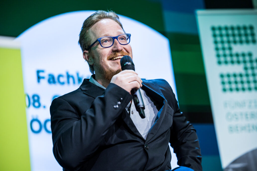 Patrick Berger vom Chancen-Nutzen-Büro des ÖGB spricht bei der Fachkonferenz des Österreichischen Behindertenrats 2026 auf der Bühne. Er sitzt im Rollstuhl, trägt einen dunklen Anzug und eine blaue Brille und hält ein Mikrofon in der Hand.