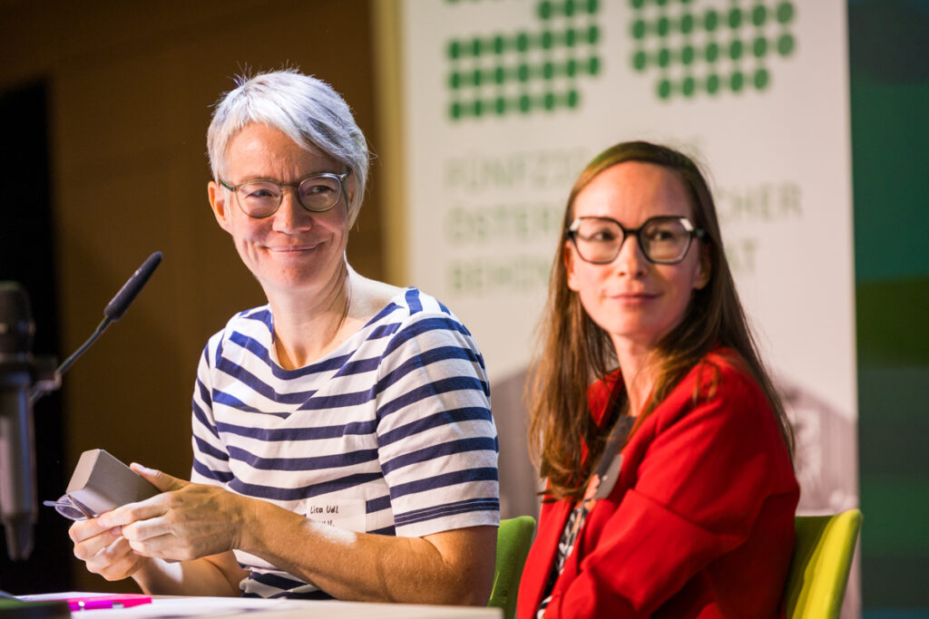 Lisa Udl (links) und Isabell Naronigg von Ninlil bei ider Fachkonferenz des Österreichischen Behindertenrats 2026. Beide sitzen auf der Bühne, Lisa Udl lächelt und hält ein Brillenetui in der Hand.