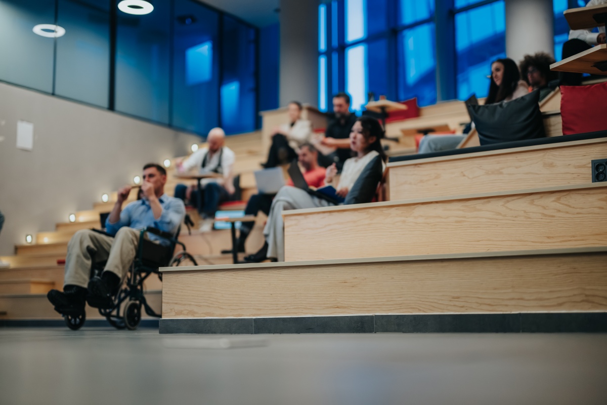 Eine vielfältige Gruppe von Studierenden, darunter eine Person im Rollstuhl, hört in einem modernen Seminarraum zu. Die Studierenden sitzen in entspannter Atmosphäre auf abgestuften Holzsitzen; vor ihnen liegen Laptops und Notizbücher.