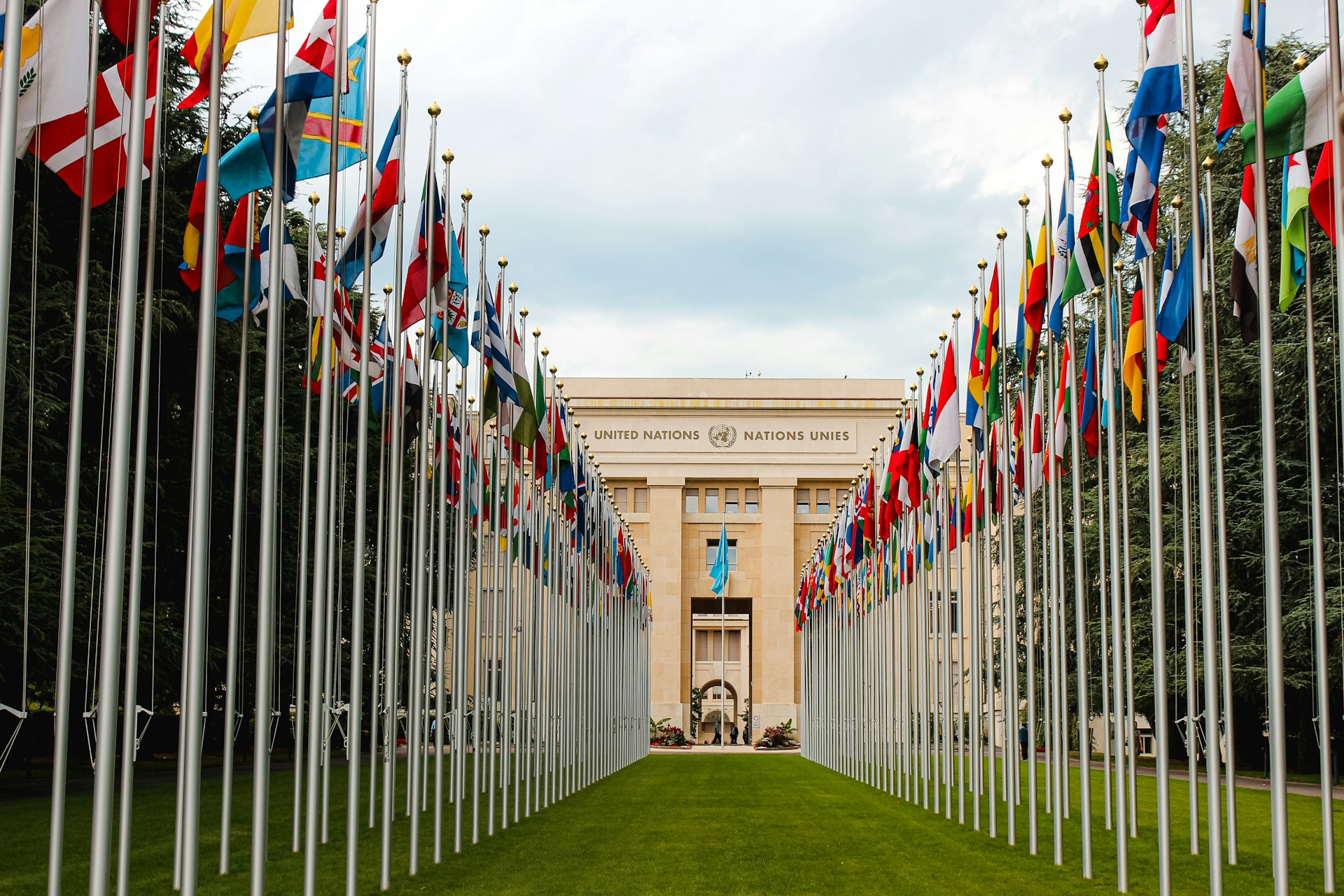Zahlreiche Nationalflaggen säumen den Weg zum Palais des Nations, dem Sitz der Vereinten Nationen in Genf. Im Hintergrund ist das Gebäude mit der Aufschrift „United Nations – Nations Unies“ zu sehen.