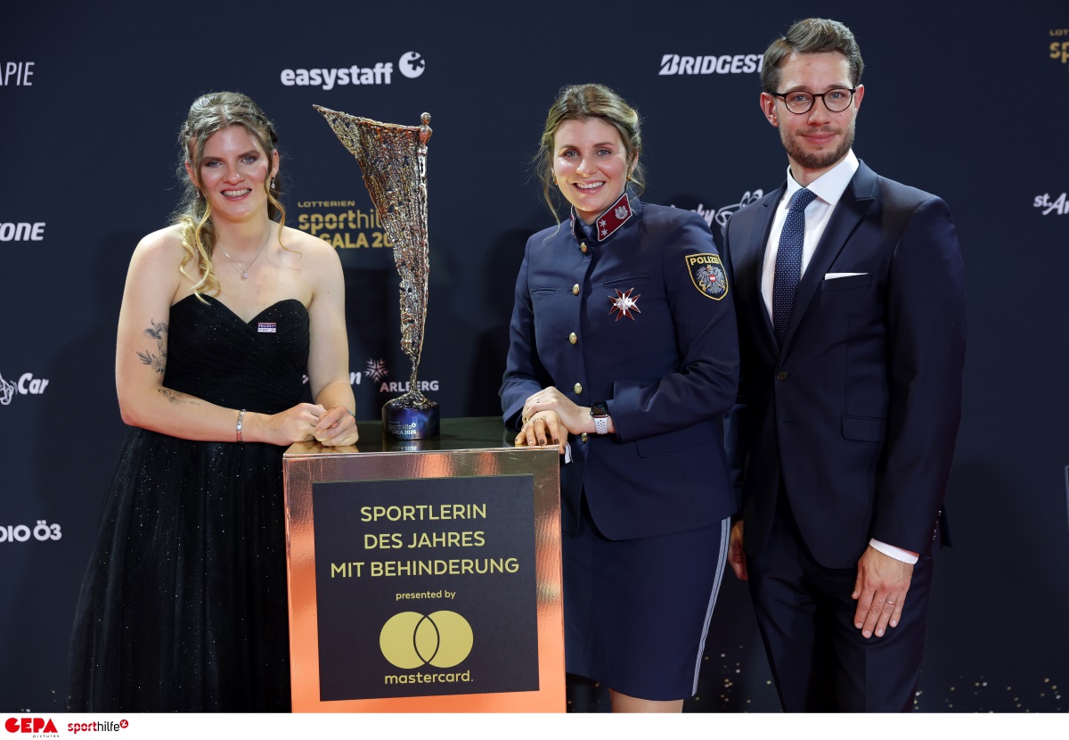 Veronika Aigner im schwarzen Abendkleid, Elisabeth Aigner in Polizeiuniform und Michael Brönner im dunklen Anzug stehen bei der Sporthilfe-Gala hinter der Auszeichnung „Sportlerin des Jahres mit Behinderung“. Zwischen ihnen steht die NIKI-Trophäe.
