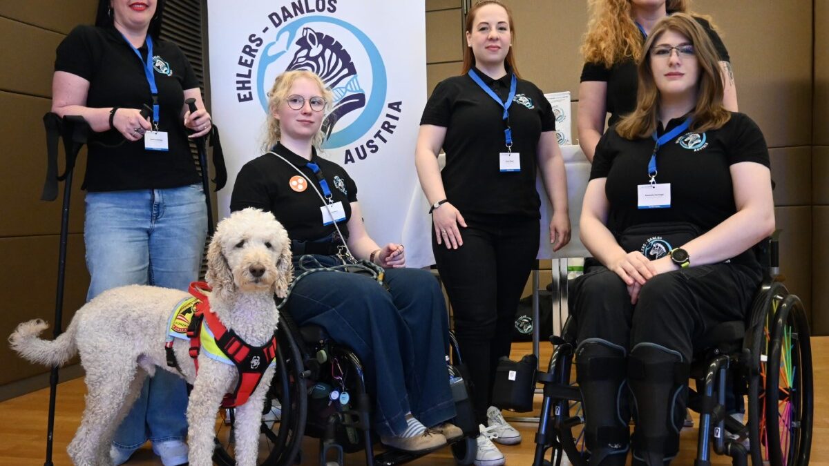 Fünf Frauen der Ehlers-Danlos Community Austria, zwei davon im Rollstuhl, posieren gemeinsam mit einem Assistenzhund vor einem Roll-up des Vereins.