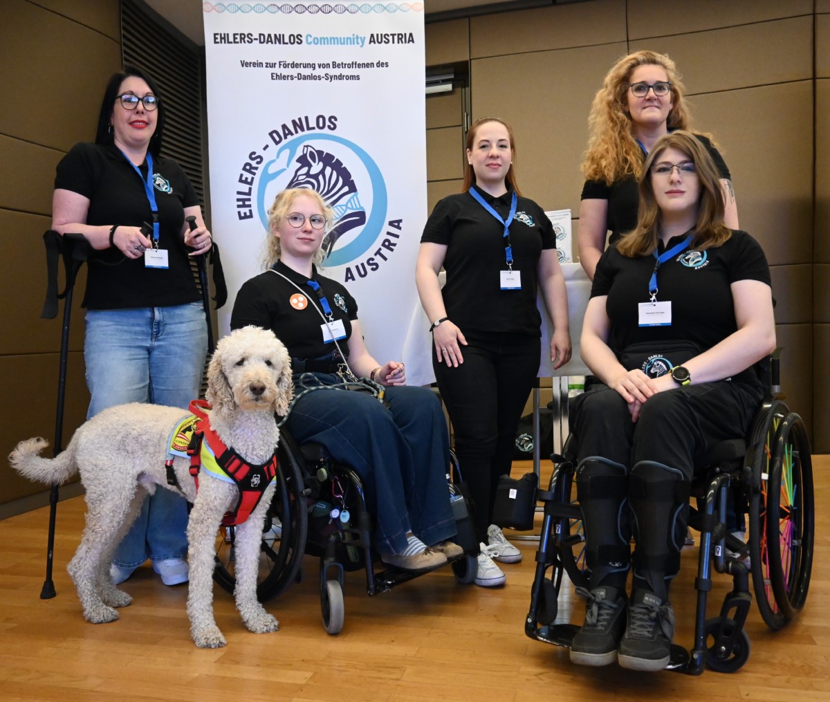 Fünf Frauen der Ehlers-Danlos Community Austria, zwei davon im Rollstuhl, posieren gemeinsam mit einem Assistenzhund vor einem Roll-up des Vereins.