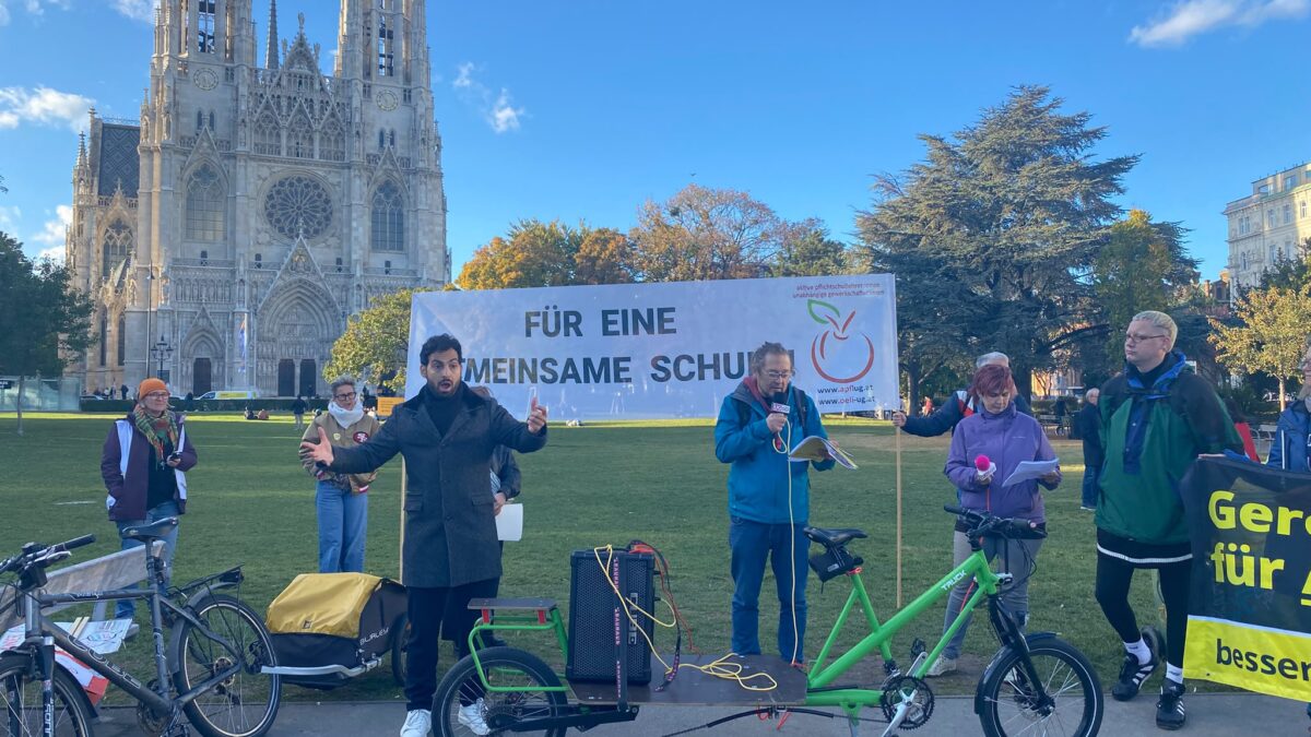 Demonstration für inklusive Bildung am 22. Oktober 2026 vor der Votivkirche in Wien. Im Vordergrund übersetzt Deilil Yilmaz in schwarzem Mantel eine Rede in Österreichische Gebärdensprache (ÖGS). Neben ihm spricht ein Mann in blauer Jacke in ein Mikrofon. Hinter ihnen halten Demonstrierende ein großes Transparent mit der Aufschrift „Für eine gemeinsame Schule“. Rechts ist ein weiteres Transparent mit der Forderung nach gerechter Bildung zu sehen. Vor der Gruppe steht ein grünes Lastenrad mit Lautsprecheranlage.