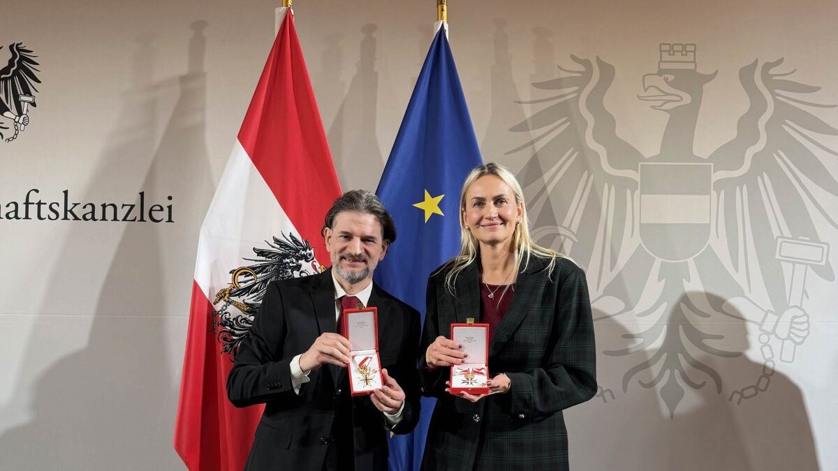 Helene Jarmer und Lukas Huber zeigen ihre staatlichen Auszeichnungen in geöffneten Etuis. Im Hintergrund sind die österreichische und die EU-Flagge sowie das Staatswappen zu sehen.
