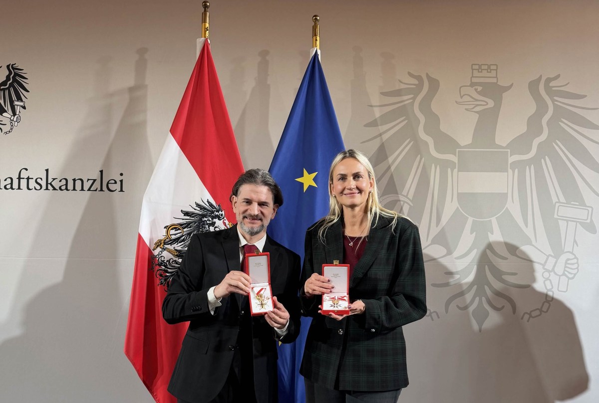 Helene Jarmer und Lukas Huber zeigen ihre staatlichen Auszeichnungen in geöffneten Etuis. Im Hintergrund sind die österreichische und die EU-Flagge sowie das Staatswappen zu sehen.