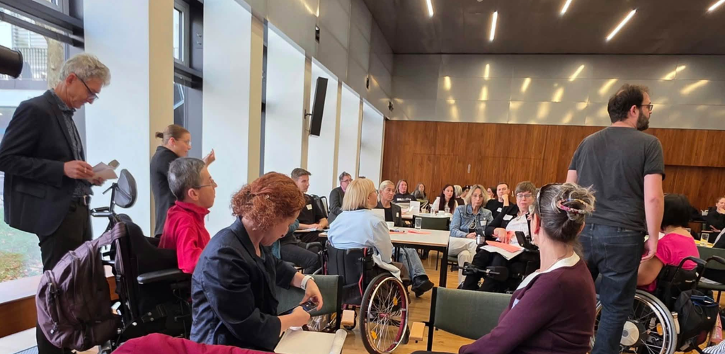 Teilnehmende des Dialogforums zu Persönlicher Assistenz von LICHT INS DUNKEL in St. Pölten sitzen an mehreren Tischgruppen und diskutieren miteinander. Einige Personen nutzen Rollstühle. Im Vordergrund sitzen mehrere Teilnehmende, während ein Mann rechts im Bild steht und zur Gruppe spricht.