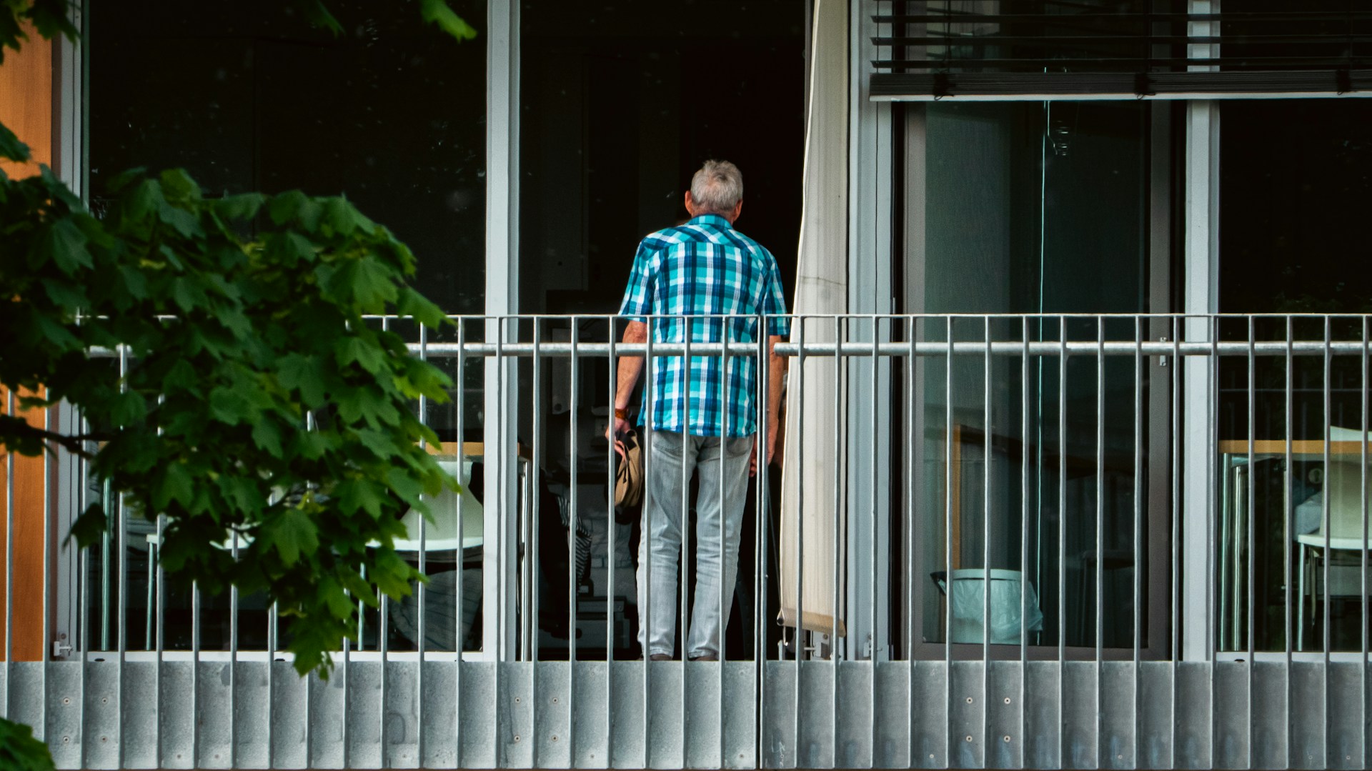 Ein älterer Mann mit grauen Haaren und blau kariertem Hemd steht mit dem Rücken zur Kamera auf einem Balkon vor einer geöffneten Glastür. Im Vordergrund sind ein Metallgeländer und grüne Baumblätter zu sehen.