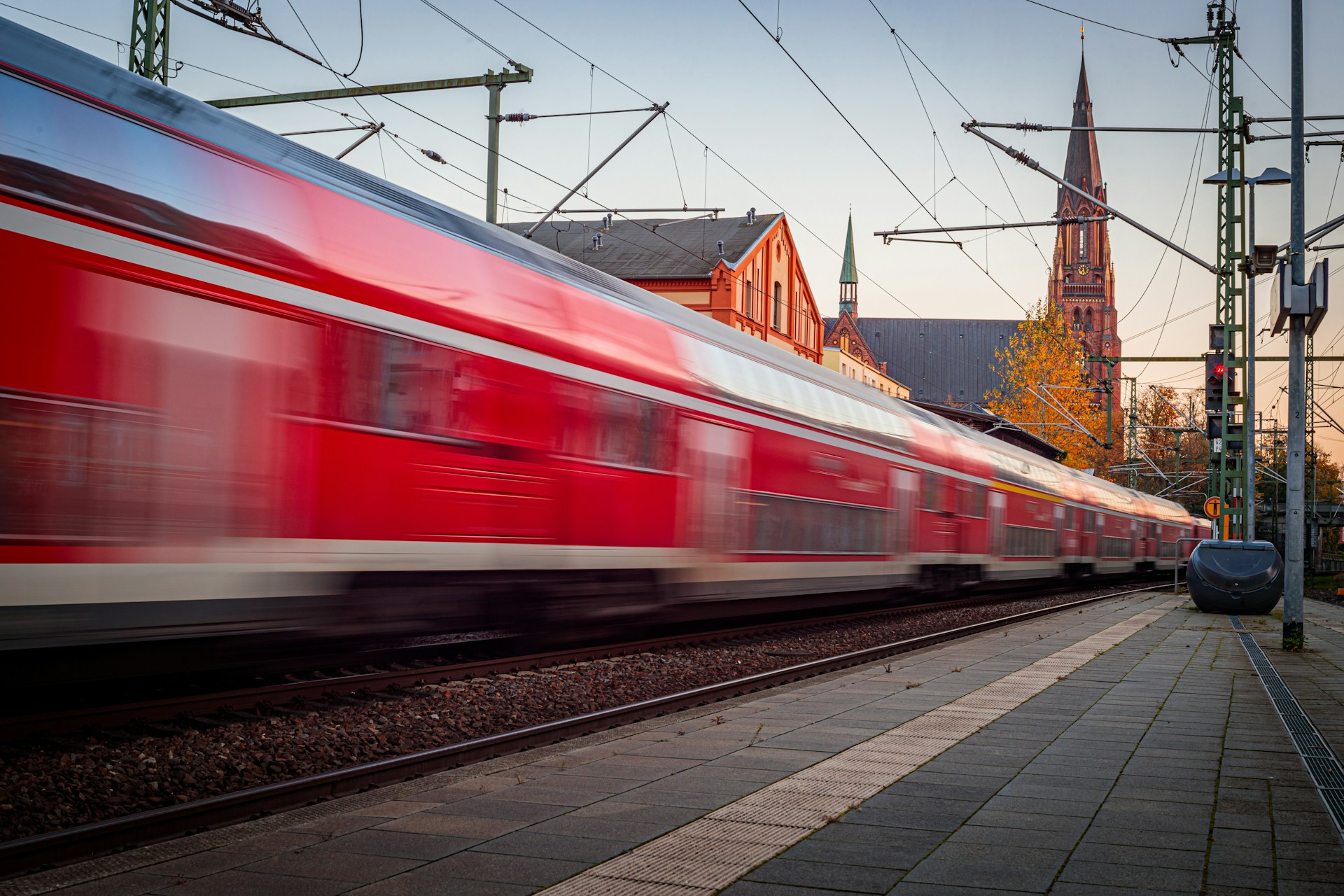 Ein rot-weißer Personenzug fährt mit hoher Geschwindigkeit an einem Bahnsteig vorbei. Im Hintergrund sind Bahnhofsgebäude, Oberleitungen und ein hoher Kirchturm im Abendlicht zu sehen.